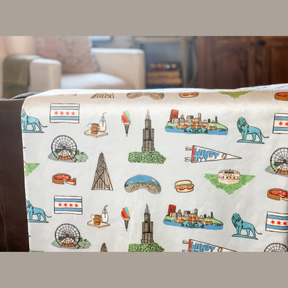 Cozy Chicago-themed plush throw blanket, 60x80 inches, featuring iconic city landmarks like deep dish, a Ferris wheel, lion, bean, a boat on a green river, Windy City flag, and other elements in vibrant colors.