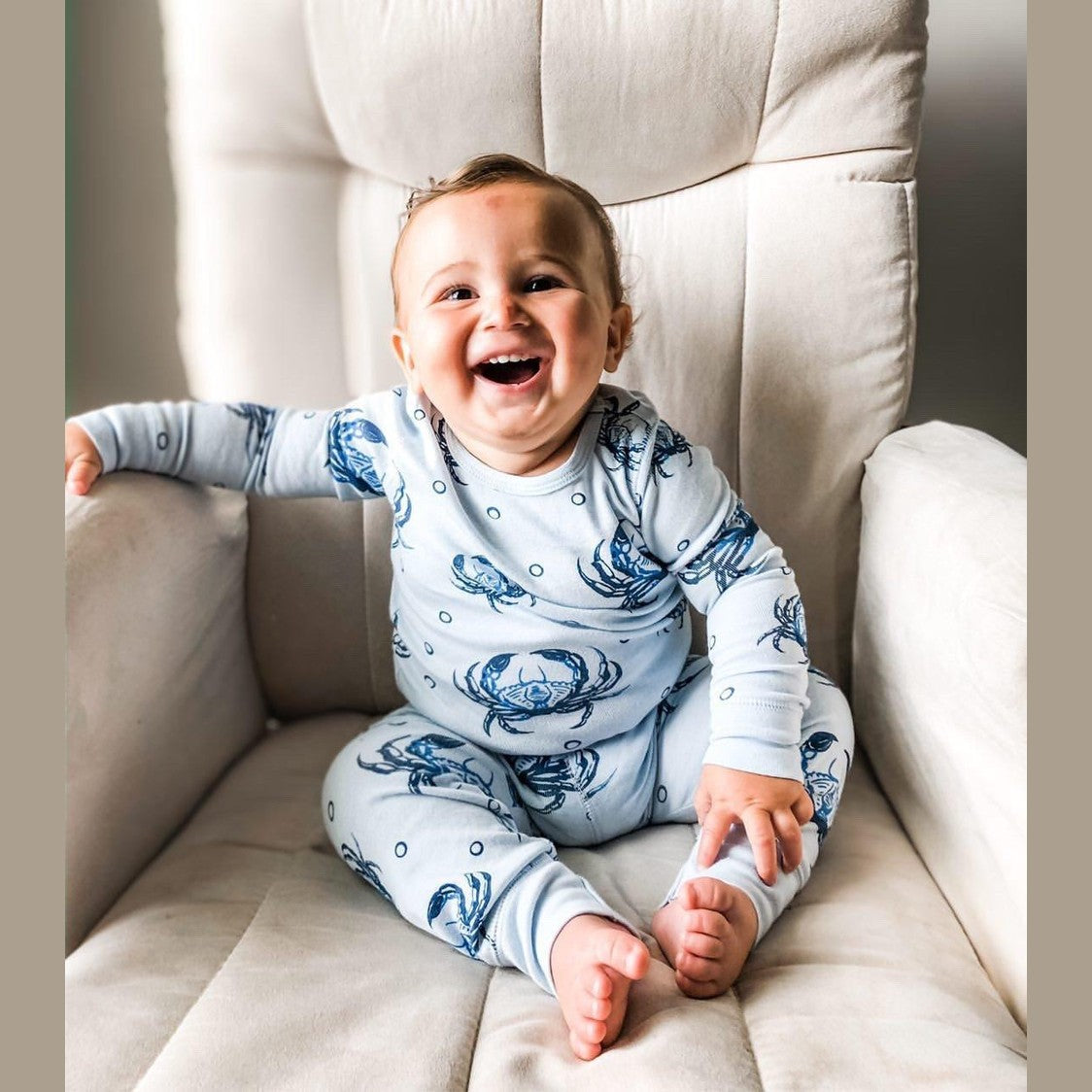 Toddler wearing blue pajamas with playful crab patterns, smiling and holding a stuffed animal in a cozy room.