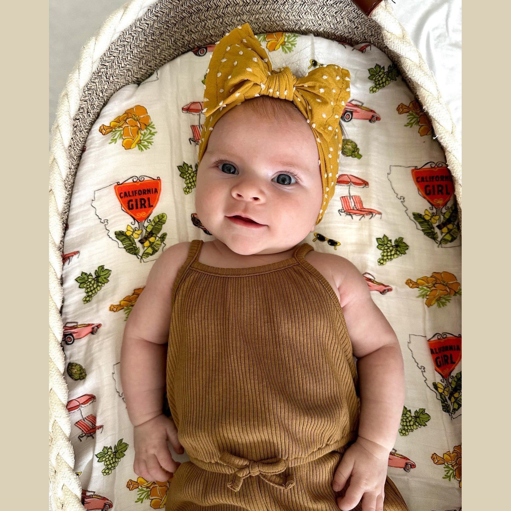 Baby girl wrapped in a soft, white muslin swaddle blanket with "California" and colorful icons printed on it.