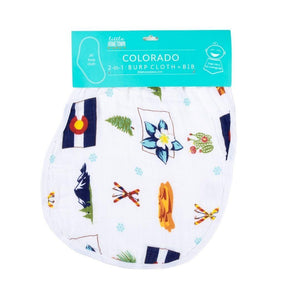 Smiling baby wearing a colorful bib with Colorado-themed illustrations, lying on a soft white blanket.