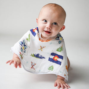 Smiling baby wearing a colorful bib with a mountain design, lying on a soft, white blanket.
