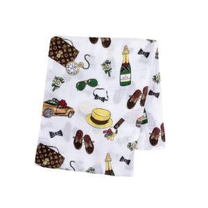 Baby swaddled in a white muslin blanket with blue and green dapper animal prints, lying on a soft surface.