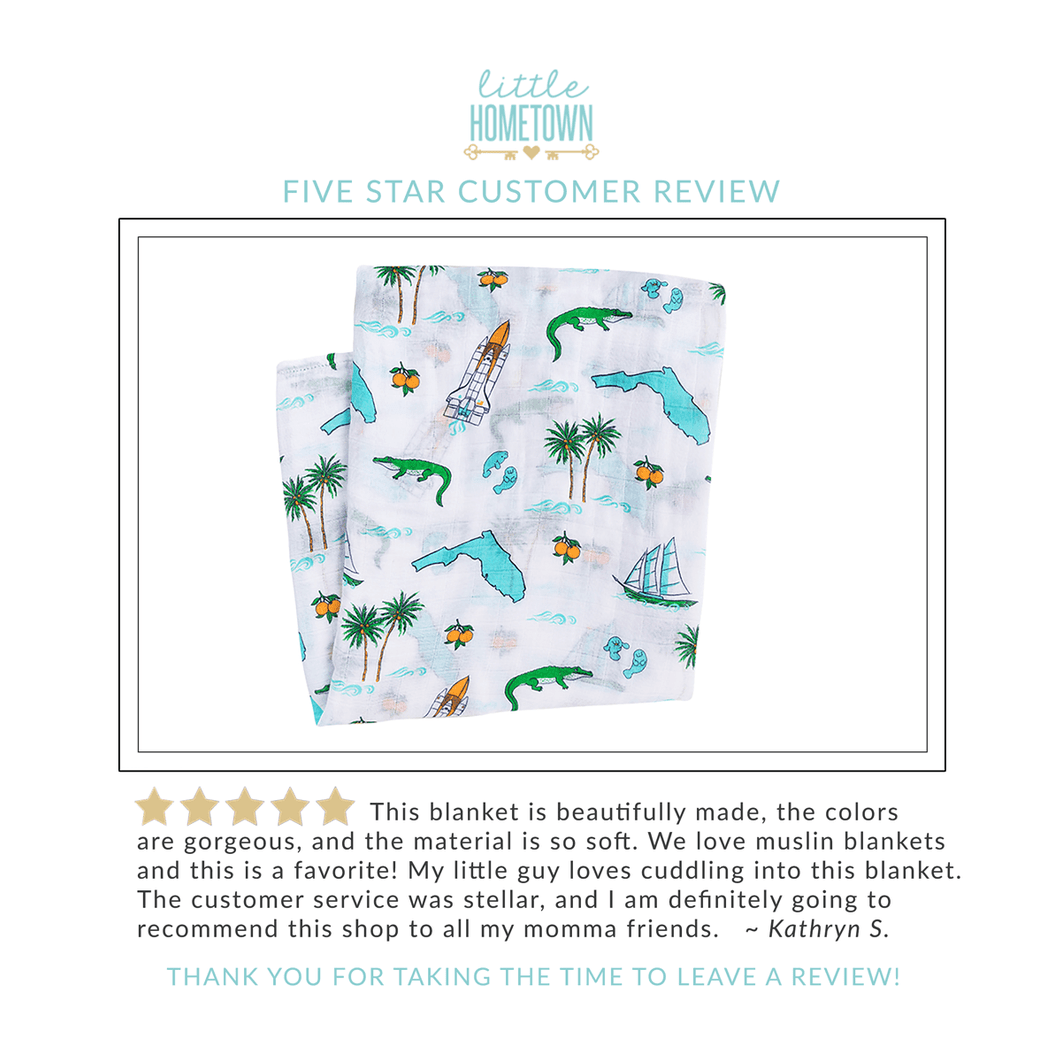 White muslin swaddle blanket with colorful Florida-themed illustrations, including oranges, flamingos, and palm trees.