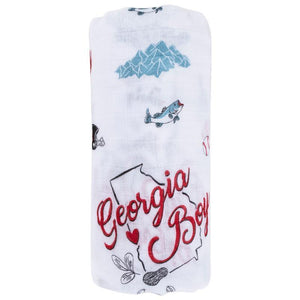 Baby swaddled in a white muslin blanket with "Georgia Boy" text and peach illustrations, lying on a soft surface.