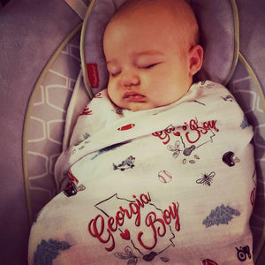 Baby swaddled in a white muslin blanket with "Georgia Boy" text and peach illustrations, lying on a soft surface.