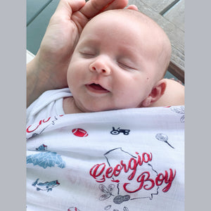 Baby swaddled in a white muslin blanket with "Georgia Boy" text and peach illustrations, lying on a soft surface.