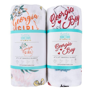 Baby swaddled in a white muslin blanket with "Georgia Boy" text and peach illustrations, lying on a soft surface.