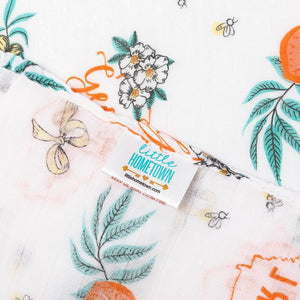 Baby swaddled in a white muslin blanket with "Georgia Girl" text and peach illustrations, lying on a soft surface.