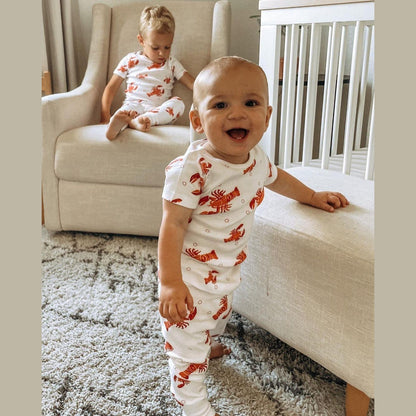 Toddler wearing white pajamas with red lobsters and crawfish, smiling and sitting on a white bed.