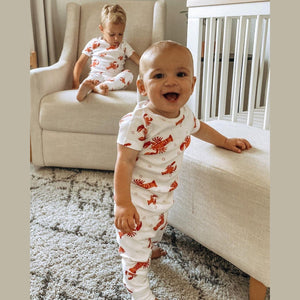 Toddler wearing white pajamas with red lobsters and crawfish, smiling and sitting on a white bed.
