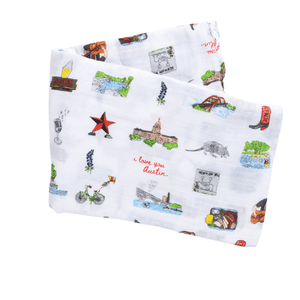 White muslin baby swaddle blanket with colorful Austin-themed illustrations, including landmarks and icons.