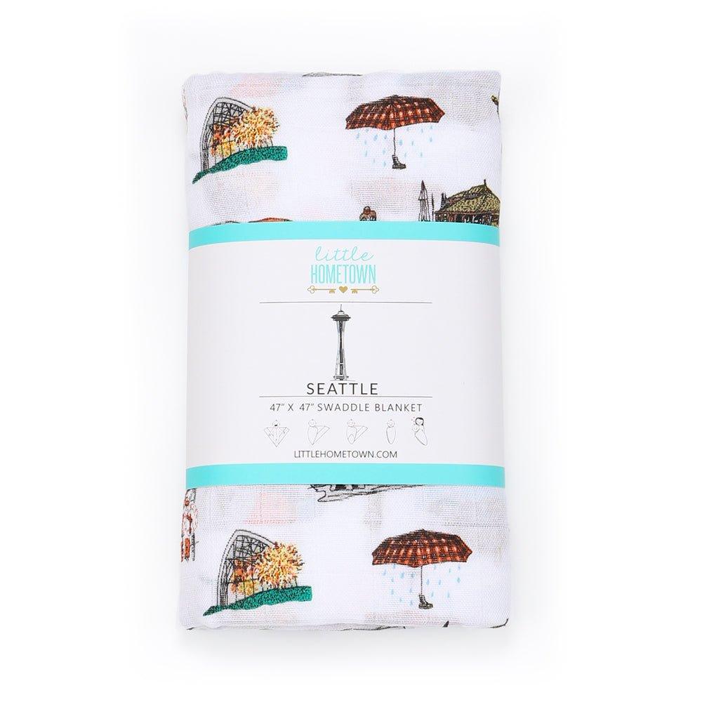 White muslin swaddle blanket with Seattle skyline, Space Needle, and Mount Rainier illustrations in soft pastel colors.