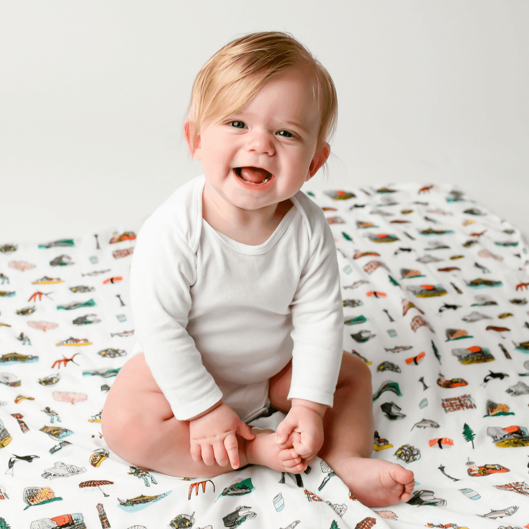White muslin swaddle blanket with Seattle skyline, Space Needle, and Mount Rainier illustrations in soft pastel colors.