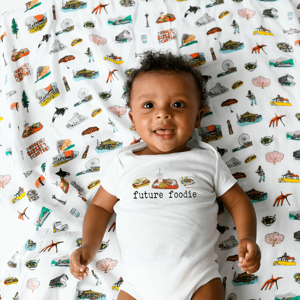 White baby onesie with "Seattle Foodie" text and icons of a coffee cup, fish, and Space Needle.
