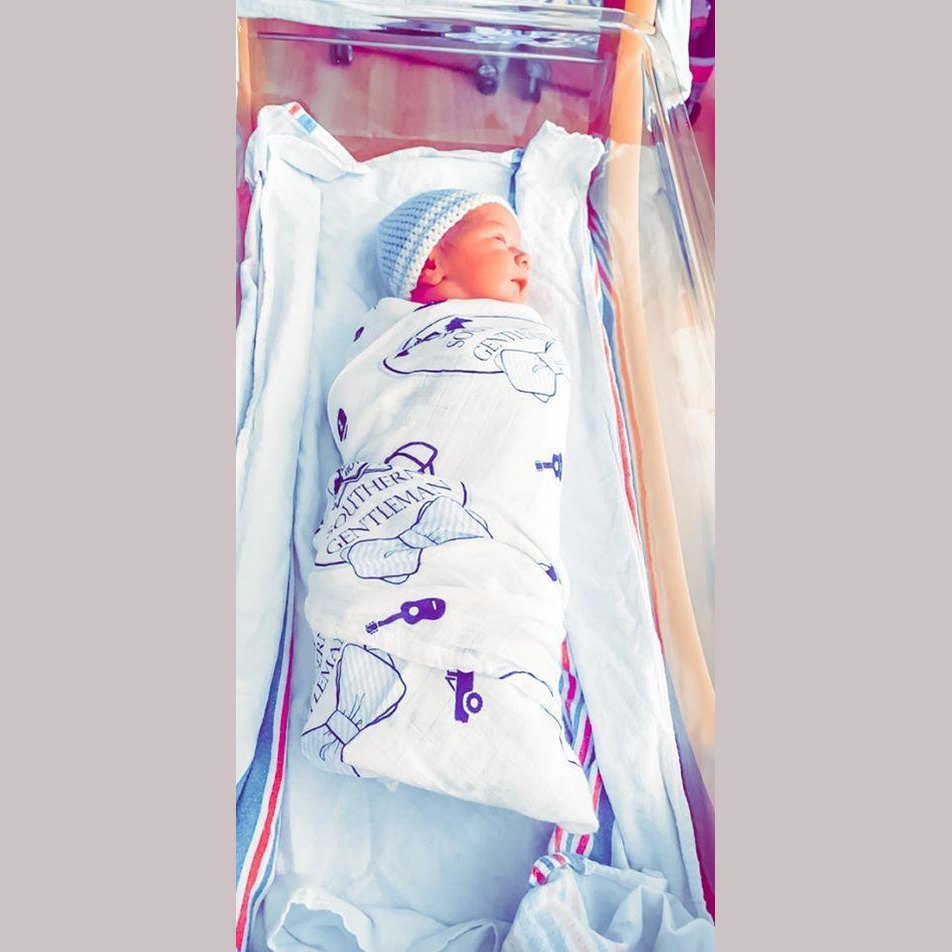 Baby boy swaddled in a white muslin blanket with blue bow ties and mustaches, lying on a soft surface.