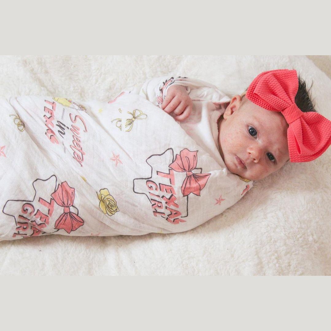 Baby girl swaddled in a white muslin blanket with pink Texas-themed illustrations, including cacti and cowboy boots.
