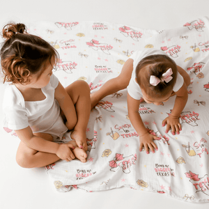 Texas-themed baby girl muslin swaddle blanket with pink accents, featuring state icons and "Texas Baby Girl" text.