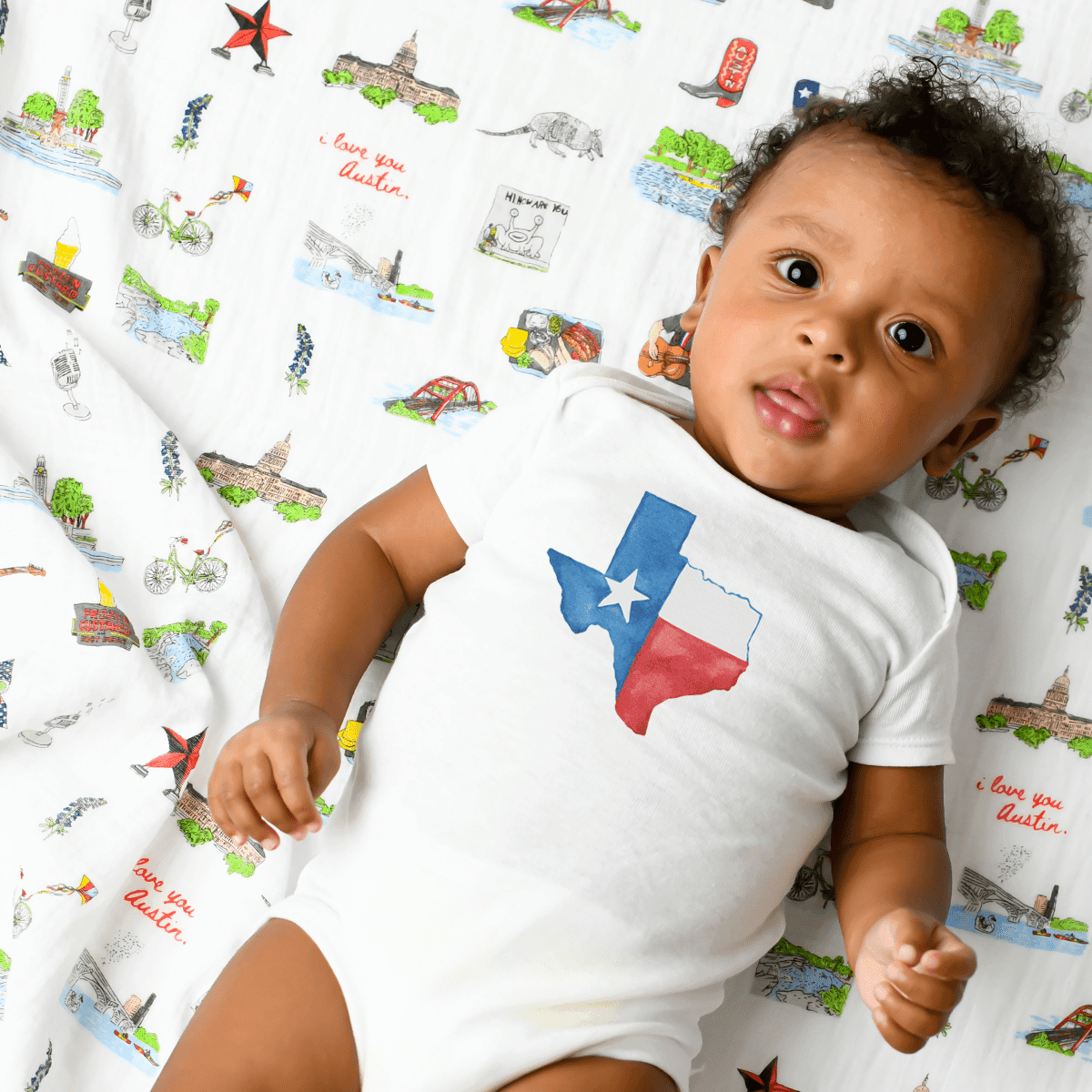 White baby onesie with a watercolor Texas map in blue and green, featuring the text "Texas" in cursive.
